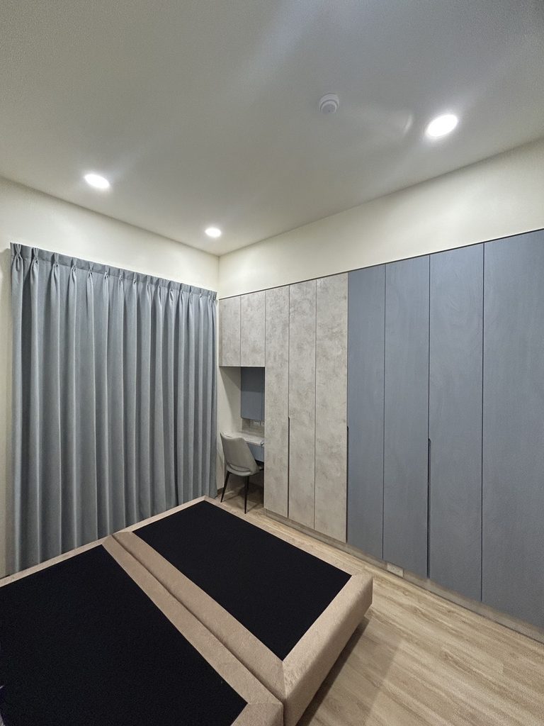 新北市土城區|光影與私密的彈性設計,客廳、主臥、客書房的布簾、風琴簾與電動捲簾全域規劃 9 由全景視角可見,中性灰色對開布簾成為空間背景的穩定基調,與暖米黃色牆面、溫潤木紋地板,以及側面帶有些微莫蘭迪質感的灰藍色系統櫃互相串聯,營造出安穩沉靜的閱讀與工作氛圍。